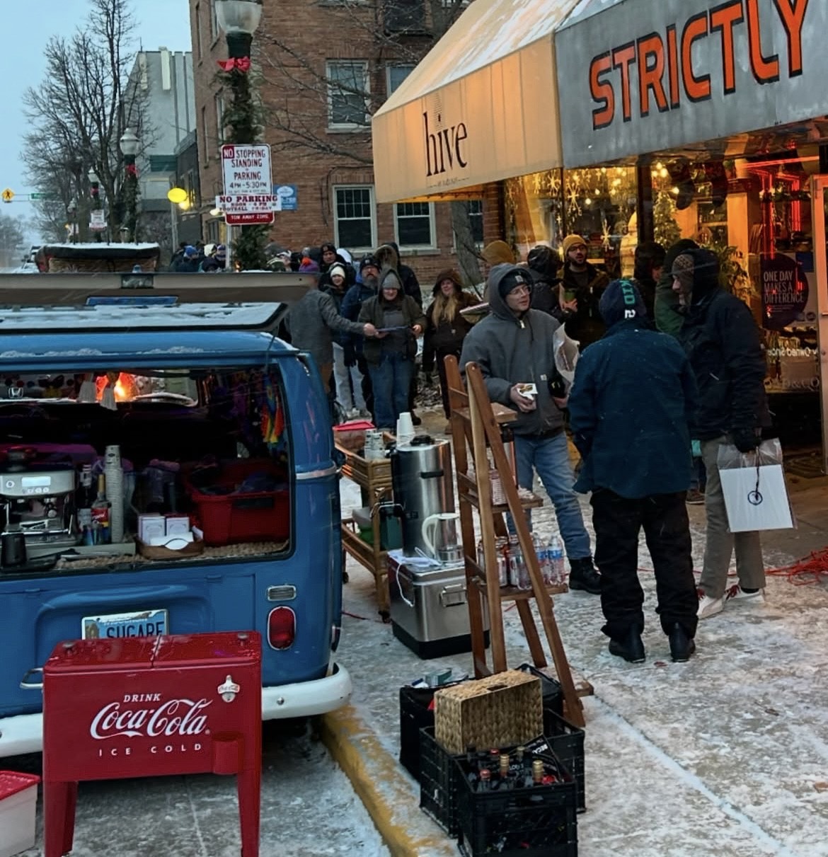 People gathered around a blue van with a Coca-Cola cooler on a snowy street, near a building with 'Strictly Hive' sign.